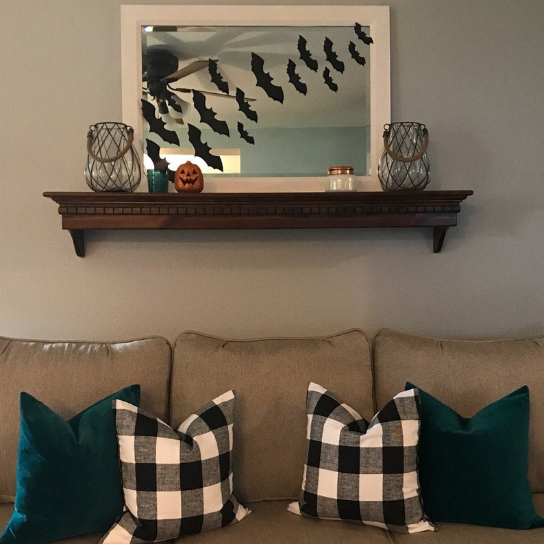 May include: A living room decorated for Halloween. A dark wooden shelf holds decorative items, including two glass lanterns, a small pumpkin, and a candle. Black bat cutouts are attached to the mirror above. The couch has green and black and white checkered pillows.
