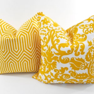 May include: Two decorative throw pillows with yellow and white patterns. One pillow features a geometric design, while the other has a floral and scrollwork pattern. The pillows are square and appear to be made of fabric.
