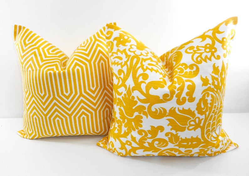 May include: Two decorative throw pillows with yellow and white patterns. One pillow features a geometric design, while the other has a floral and scrollwork pattern. The pillows are square and appear to be made of fabric.