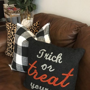 May include: A collection of decorative throw pillows on a brown leather couch. Pillows include a black and white buffalo check, leopard print, and a black pillow with the words "Trick or Treat" embroidered in orange and gray.