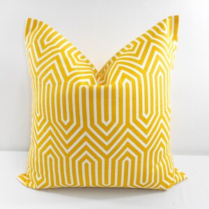 May include: A square decorative pillow with a geometric pattern. The pillow features a bright yellow background with a white, repeating, angular design. The pillow is placed on a white surface.