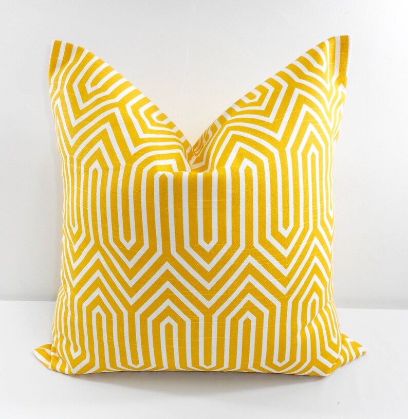 May include: A square decorative pillow with a geometric pattern. The pillow features a bright yellow background with a white, repeating, angular design. The pillow is placed on a white surface.