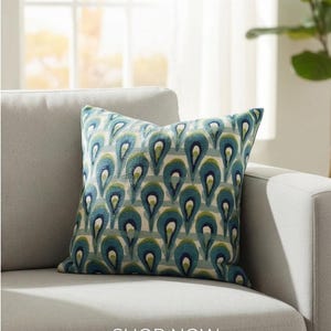 Pu&ograve; includere: Un cuscino quadrato decorativo con un motivo a goccia turchese e blu su sfondo chiaro. Il cuscino &egrave; su un divano grigio. Il testo "SHOP NOW" &egrave; in basso nell'immagine.
