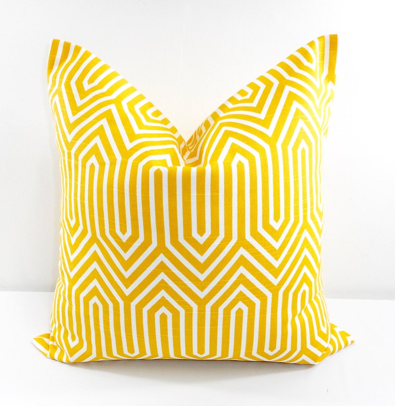 May include: A square decorative pillow with a geometric pattern. The pillow features a repeating design of white lines on a yellow background. The pillow is made of fabric and is likely used for home decor.