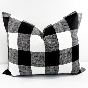 May include: A square decorative pillow with a black and white buffalo check pattern. The pillow has a textured appearance and is designed for home decor. The pillow is a neutral color and can be used in a variety of settings.