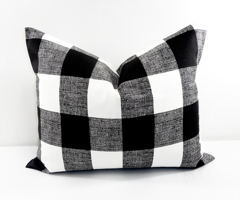 May include: A square decorative pillow with a black and white buffalo check pattern. The pillow has a textured appearance and is designed for home decor. The pillow is a neutral color and can be used in a variety of settings.