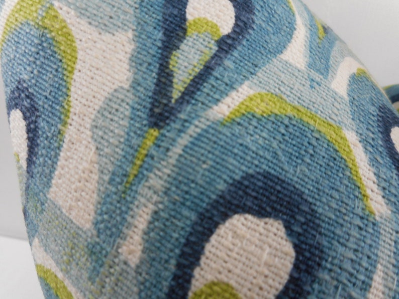Pu&ograve; includere: Primo piano di un tessuto strutturato con un motivo ripetuto di forme organiche blu, turchesi, bianche e verde lime. Il tessuto ha un aspetto tessuto, che suggerisce un cuscino o un materiale di rivestimento.