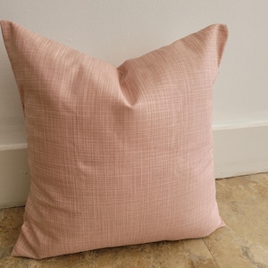 Puede incluir: Una almohada de sofá suave de color rosa claro con un tejido texturizado sutil. La almohada es cuadrada y tiene un diseño simple y moderno.