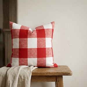 May include: A square decorative pillow with a red and white gingham pattern. The pillow is placed on a wooden bench with a neutral-colored fabric draped over it. The pillow's design features large, evenly sized checks.