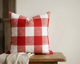 May include: A square decorative pillow with a red and white gingham pattern. The pillow is placed on a wooden bench with a neutral-colored fabric draped over it. The pillow's design features large, evenly sized checks.