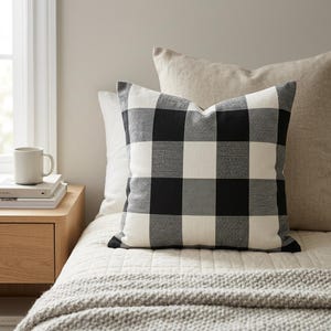 May include: A black and white buffalo check pattern pillow rests on a bed with other pillows. The pillow has a square shape and is made of a soft fabric. The bed is covered with a white quilt and a gray blanket.