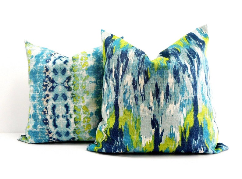 Pu&ograve; includere: Due cuscini decorativi quadrati con motivi astratti. Uno presenta un design tie-dye blu e bianco, l'altro una miscela di pennellate blu, verdi e bianche. I cuscini hanno un aspetto morbido e strutturato.