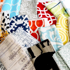 May include: A collection of fabric swatches in various colours and patterns, including orange, yellow, blue, red, and green. The fabrics feature geometric, floral, and abstract designs. Some of the fabrics have text printed on them, including "1889", "CARTE POSTALE", and "GRANDE MAISON DE PARIS".