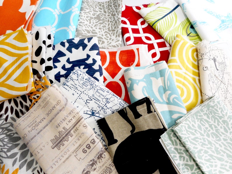 May include: A collection of fabric swatches in various colours and patterns, including orange, yellow, blue, red, and green. The fabrics feature geometric, floral, and abstract designs. Some of the fabrics have text printed on them, including "1889", "CARTE POSTALE", and "GRANDE MAISON DE PARIS".