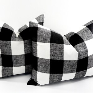 May include: Two square throw pillows with a black and white buffalo check pattern. The pillows are made of a textured fabric and are displayed against a white background. The pillows are a decorative home decor item.