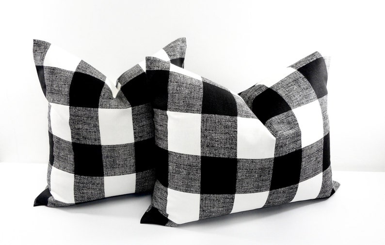 May include: Two square throw pillows with a black and white buffalo check pattern. The pillows are made of a textured fabric and are displayed against a white background. The pillows are a decorative home decor item.