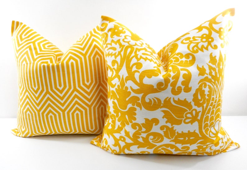 May include: Two decorative throw pillows with yellow and white patterns. One pillow features a geometric design, while the other showcases a floral damask pattern. The pillows are square and appear to be made of fabric.