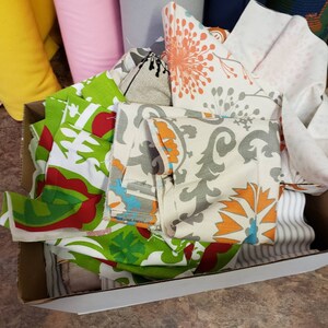 May include: A white box filled with colourful fabric scraps. The fabrics have various patterns, including floral, geometric, and striped designs. The fabrics are in shades of green, red, orange, white, and grey.