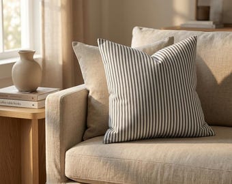 May include: A striped pillow with black and white vertical stripes rests on a beige sofa. The pillow is square and has a soft, inviting appearance. The sofa is neutral-toned, and the scene is bathed in natural light.
