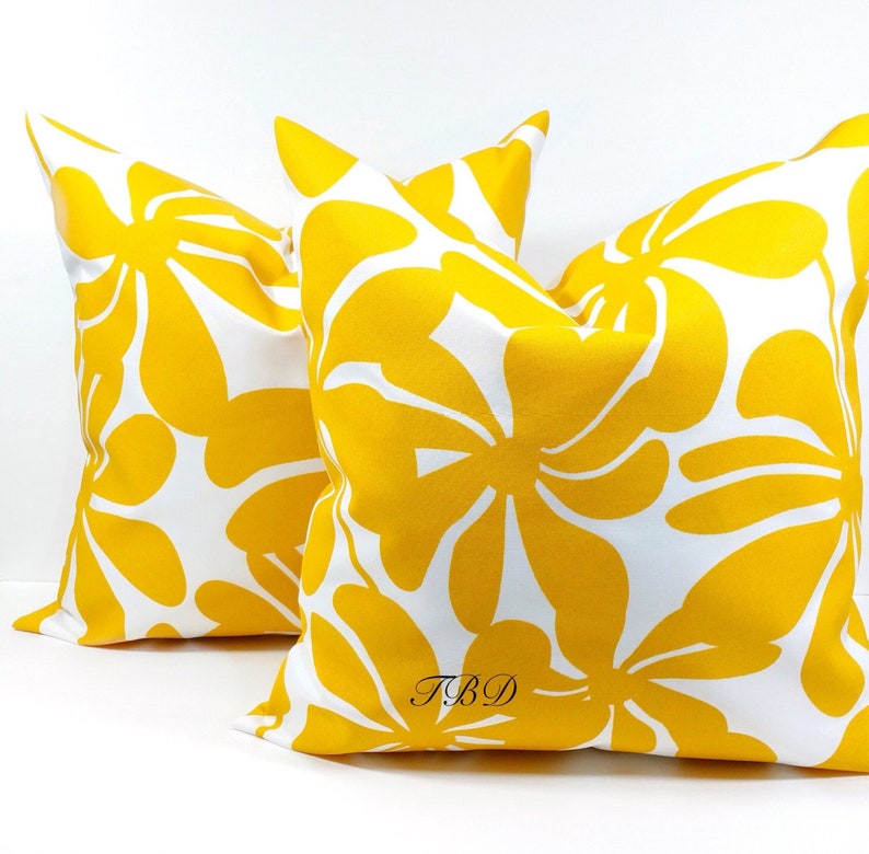May include: Two yellow throw pillows with a white background and a floral pattern. The pillows have the text "TBD" printed on them.