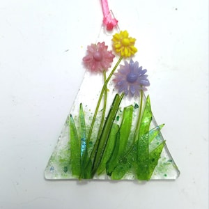 May include: A triangular glass ornament with a floral design. The ornament features three flowers in pink, yellow, and purple, with green stems and grass. A pink ribbon is attached at the top for hanging.