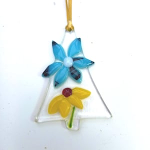 May include: A clear glass Christmas tree ornament shaped like a tree, decorated with two flower designs. The top flower is blue with black accents, and the bottom flower is yellow with a red centre. A gold ribbon is attached.
