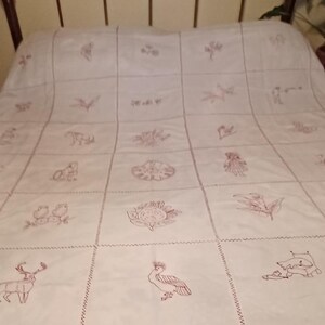 May include: White bedspread with a grid pattern, featuring embroidered red images of flowers, birds, and animals. The bedspread is on a vintage metal bed frame, creating a rustic aesthetic.