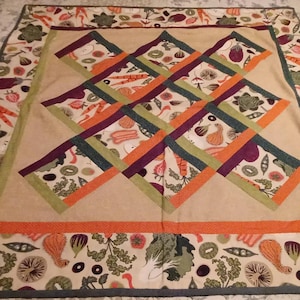 May include: A square quilt with a beige center featuring a crisscross design of orange, green, and purple stripes. The border is patterned with various vegetables and fruits, including carrots, eggplants, and strawberries.