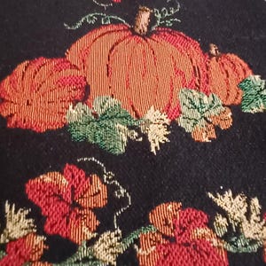 May include: Black fabric table runner with embroidered pumpkins and fall foliage in shades of orange, red, and green. The runner features a repeating pattern of pumpkins and leaves, perfect for autumn decor.