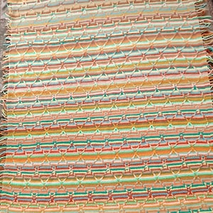 May include: A colorful, handmade crochet blanket with a geometric pattern. The blanket features horizontal stripes in shades of orange, green, yellow, and pink. The edges are finished with a green border and fringe.