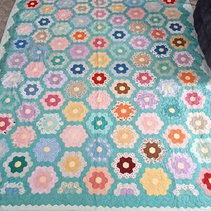 May include: A handmade quilt with a turquoise background and numerous hexagon-shaped floral designs. The flowers are in various pastel colors, including pink, yellow, and purple, with white lace accents. The quilt has a scalloped edge.