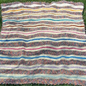 May include: A handmade crochet blanket with horizontal stripes in various colors, including white, yellow, teal, brown, pink, blue, and purple. The blanket is made with a textured stitch and has a scalloped edge. The blanket is lying on green grass.