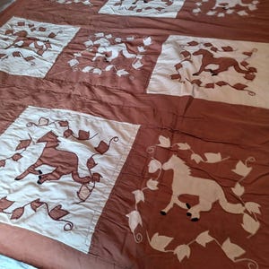 May include: A quilt featuring a repeating pattern of square panels in alternating brown and off-white colors. Each panel displays a horse silhouette surrounded by a vine and leaf border. The quilt is constructed with stitched seams.