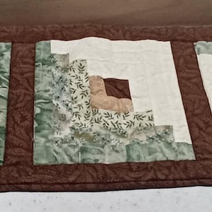 May include: A rectangular, handmade quilt with a brown border. The quilt features four square blocks with a central brown square, surrounded by cream and green patterned fabric. The quilt is likely used as a table runner.