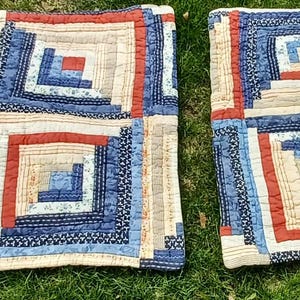 May include: Two square patchwork quilts with a log cabin design. Each quilt features a central square surrounded by concentric rectangles in shades of blue, red, and beige. The quilts are placed on green grass.
