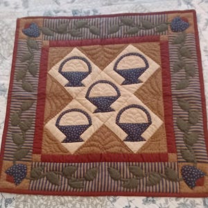 May include: A square quilted wall hanging featuring a central design of five blue baskets on a tan background. The quilt has a red border with blue and tan striped fabric and green leaf and blue flower appliqués. The overall color scheme is warm and inviting.