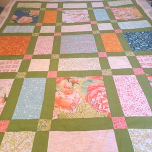 May include: A handmade quilt with a patchwork design. The quilt features rectangular blocks in various floral patterns and solid colors, including pink, orange, blue, and green, separated by green borders. The quilt is laid out on a bed.