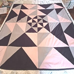 May include: A large quilt with a geometric design. The quilt features a pattern of triangles in shades of black, gray, and pink. The triangles are arranged in a symmetrical pattern, creating a starburst effect. The quilt is spread out on a bed.