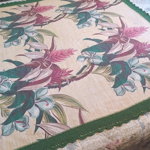May include: A square fabric tablecloth featuring a cream-colored base with a repeating floral pattern in shades of deep magenta, dark green, and light teal. The edges are finished with a dark green crocheted border with a scalloped trim.