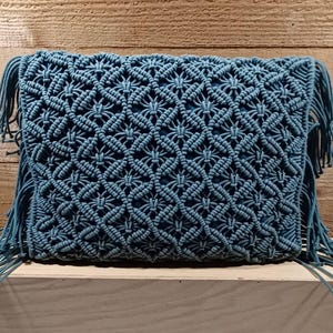 May include: A rectangular, teal macrame clutch with a geometric pattern. The clutch has fringe on the sides and is displayed on a wooden surface. The intricate knotting creates a textured, decorative design.