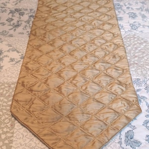 May include: A long, narrow, gold-colored table runner featuring a quilted diamond pattern across its surface. The runner has a pointed end finished with a decorative gold tassel. The fabric has a subtle sheen and is stitched with a grid design.