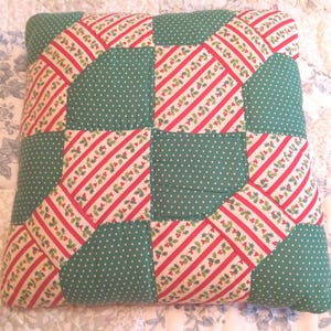 May include: A square quilted pillow featuring a patchwork design of alternating blocks. The pattern consists of green fabric with white polka dots and white fabric with diagonal red stripes and green holly leaf and red berry motifs.