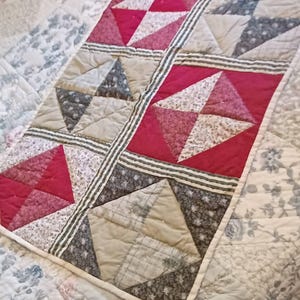 May include: A handmade quilt with a patchwork design. The quilt features squares with red, white, and gray triangles, separated by striped fabric. The background is a floral pattern in shades of gray and white.