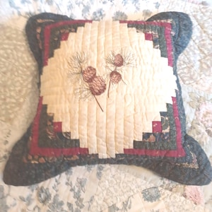 May include: A square quilted throw pillow featuring a central cream-colored panel with a stitched pinecone and needle design. The pillow is bordered by concentric layers of dark green, deep red, and a dark blue patterned fabric with a textured finish.