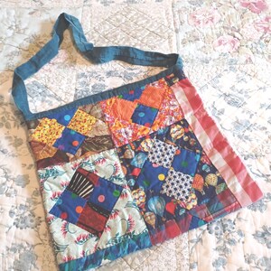 May include: A square-shaped quilted shoulder bag featuring a patchwork design of various fabric patterns including floral, geometric, and hot air balloon prints in shades of blue, orange, red, and white, with a solid teal fabric strap and trim.