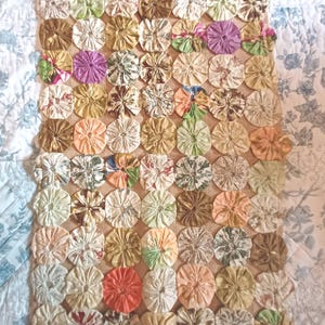 May include: A rectangular textile mat constructed from numerous gathered fabric circles, known as yo-yos, in a variety of muted colors including tan, brown, green, pink, and orange, all stitched together onto a tan burlap-like fabric backing.