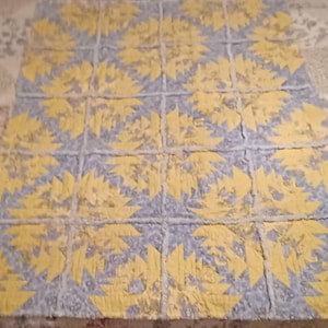 May include: A handmade quilt featuring a repeating pattern of yellow pineapple shapes on a light blue background. The quilt is made of fabric squares sewn together, with visible stitching and a textured appearance. The overall design is symmetrical and visually appealing.