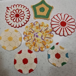 May include: A collection of seven handmade crochet coasters in various shapes and colors. The set includes circular floral designs in red and white, a pentagonal piece in green and yellow, a multi-colored flower shape, and hexagonal patterns with red, yellow, and blue accents.