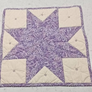 May include: A quilted star-shaped placemat. The center star is a paisley pattern in shades of purple and white. The points of the star are cream-colored squares and triangles. Each section has a small embroidered detail. The edges are bound in a coordinating fabric.