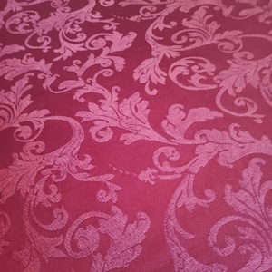 May include: A deep burgundy fabric featuring a tonal damask pattern with intricate swirling acanthus leaves and floral motifs woven into the material. The design creates a subtle texture and sheen across the surface of the dark red textile.